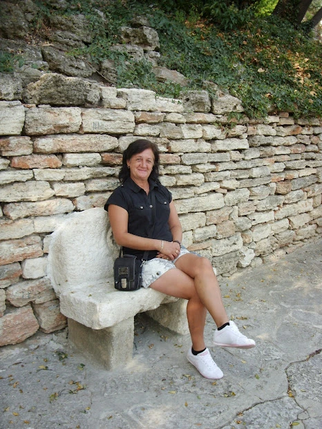 A smiling Wendy Mosdell, fibre artist, sits on a stone bench. Black shirt, denim shorts, white sneakers. Small black bag