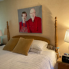 The finished Art Quilt of two people in red outfits hangs on a bedroom wall above a wooden bed with white bedding.