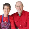 Two people sitting together, both wearing red shirts. The Woman is wearing a blue apron with a colorful pattern.